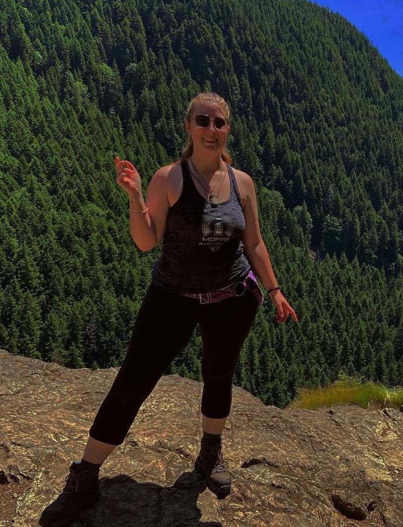 A woman stands in front of a towering mountain covered in dense coniferous trees, appearing to be on a hiking adventure