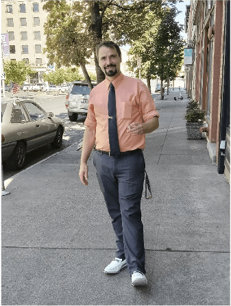 A man stands on a street, wearing a smile and holding a cigarette in his hand