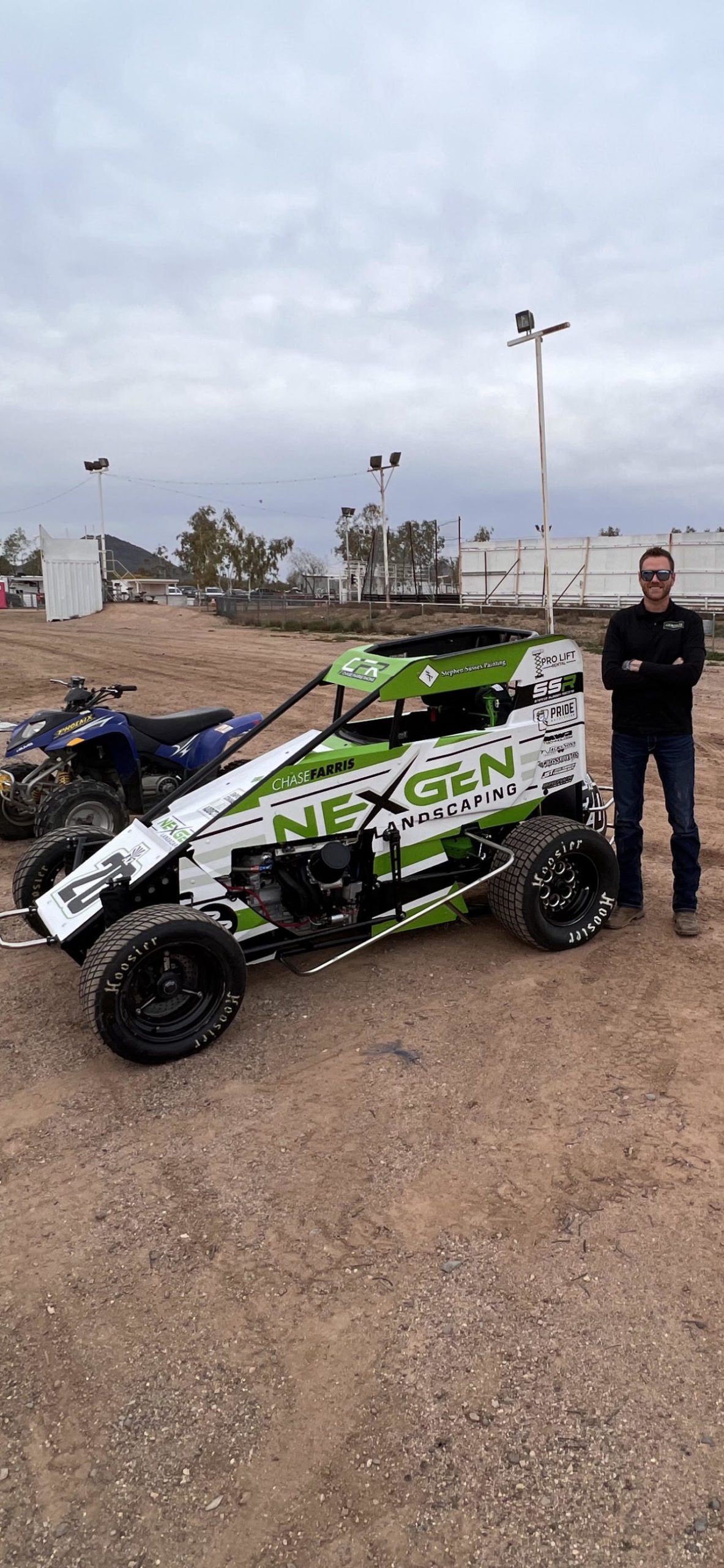 A man proudly stands beside a green sprint car with the racetrack visible in the background